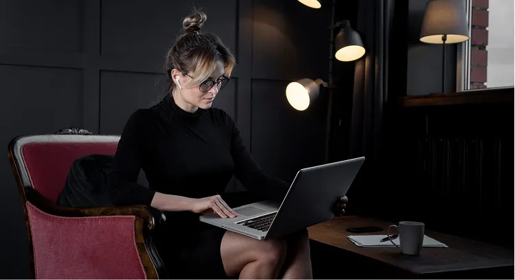 Woman in black dress sitting on a red armchair working on a laptop in a dimly lit room with lamps and a coffee cup on a table.