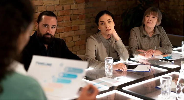 Three people seated at a table attentively listening to a presenter holding documents in an office with a brick wall background.