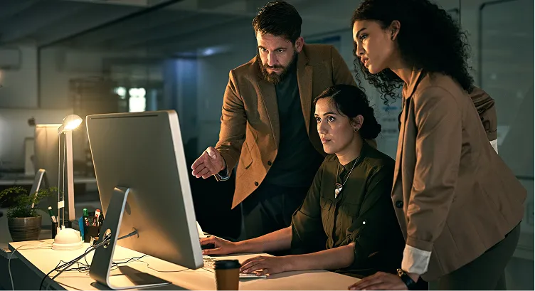 Three colleagues in business attire focused on a computer screen in a dimly lit modern office.