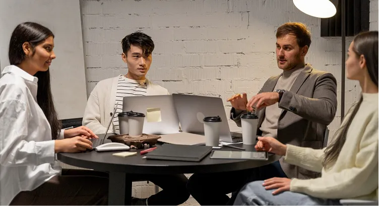 Four colleagues engaged in a meeting around a round table with laptops, coffee cups, and office supplies in a modern workspace.