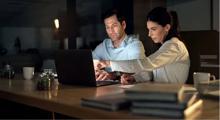 Two colleagues working late in an office, intently looking at a laptop screen with one pointing at it.