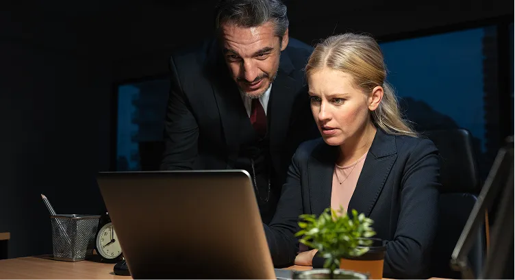 A professional man and woman working late, focused on a laptop screen in an office.