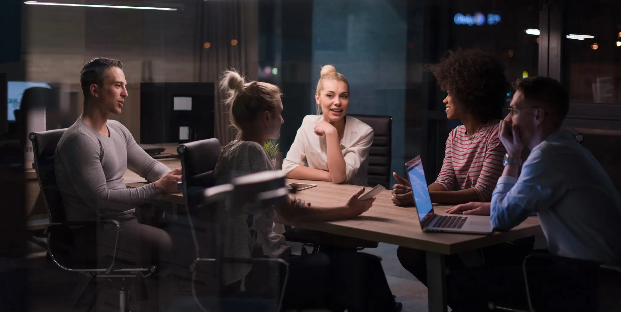 Five diverse colleagues engaged in a nighttime meeting around a conference table with laptops and smartphones.