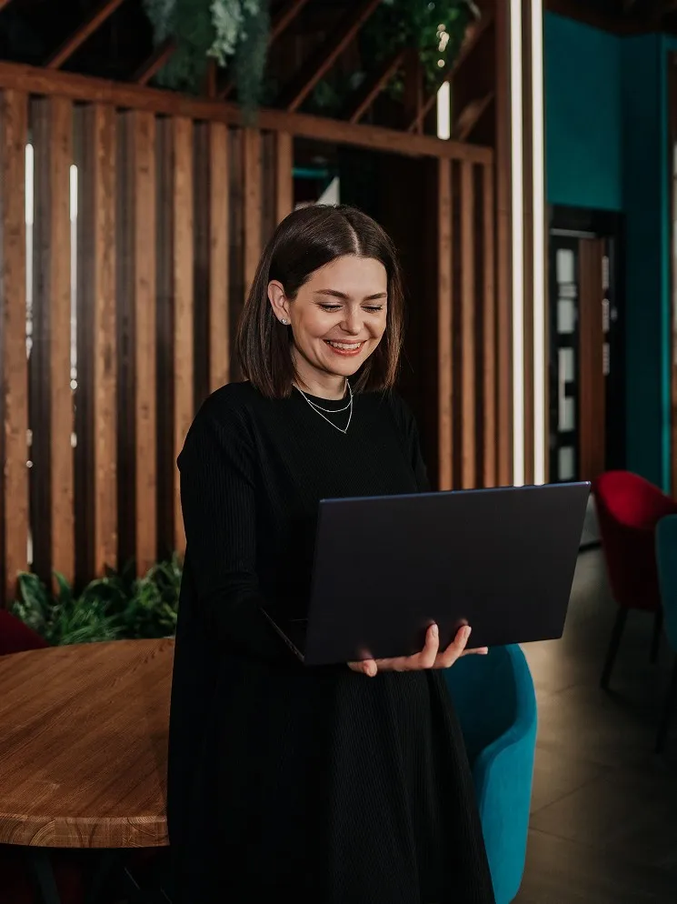Smiling woman with shoulder-length dark hair holding a laptop in a modern indoor space with wooden panels and colorful chairs.