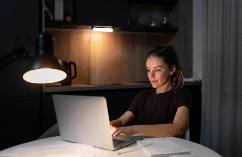 Woman working on a laptop at a table in a dimly lit room with a desk lamp.