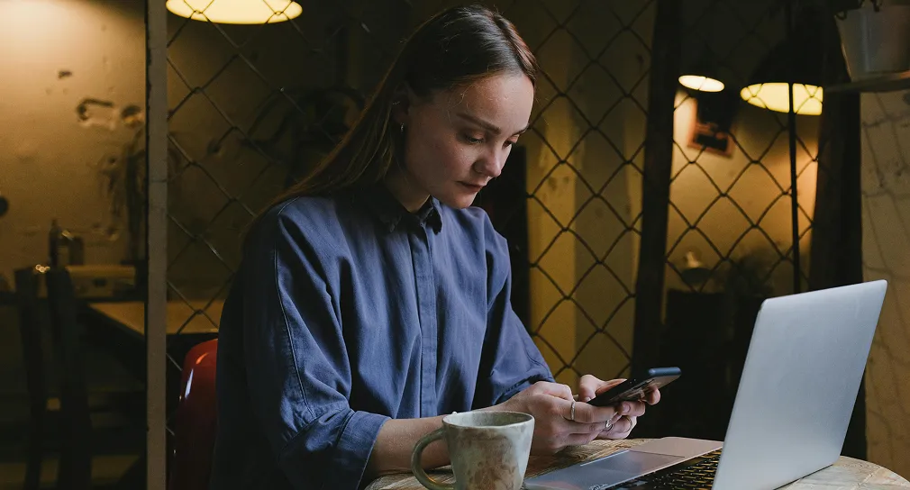 Young woman in a blue shirt sitting at a table, using a smartphone with a laptop and a coffee cup in front of her.