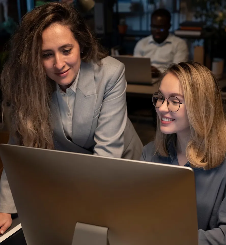 Two women working together and smiling at a computer screen in an office, with a man working on a laptop in the background.