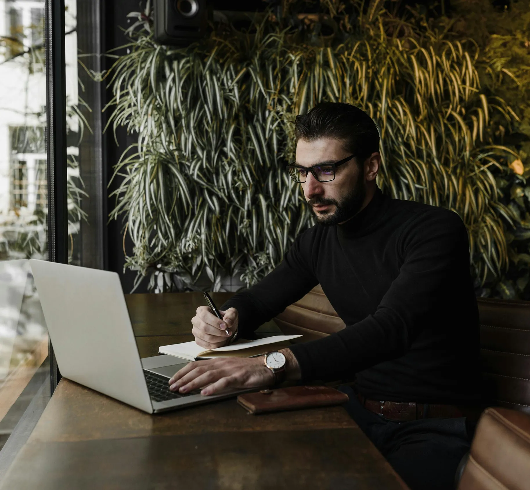 Man with glasses and black turtleneck writing in a notebook while using a laptop at a wooden table by a window with green plants in the background.