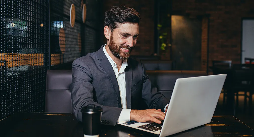 Smiling man in a suit using a laptop at a dark wooden table with a black coffee cup nearby.