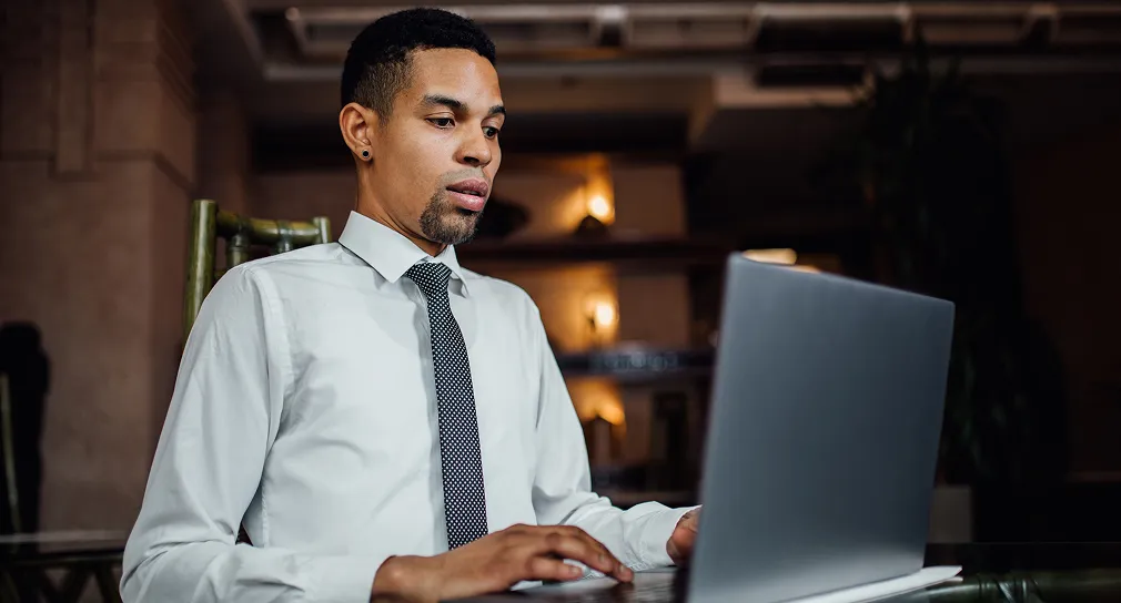 Young man in white shirt and black tie working on a laptop in a dimly lit room.