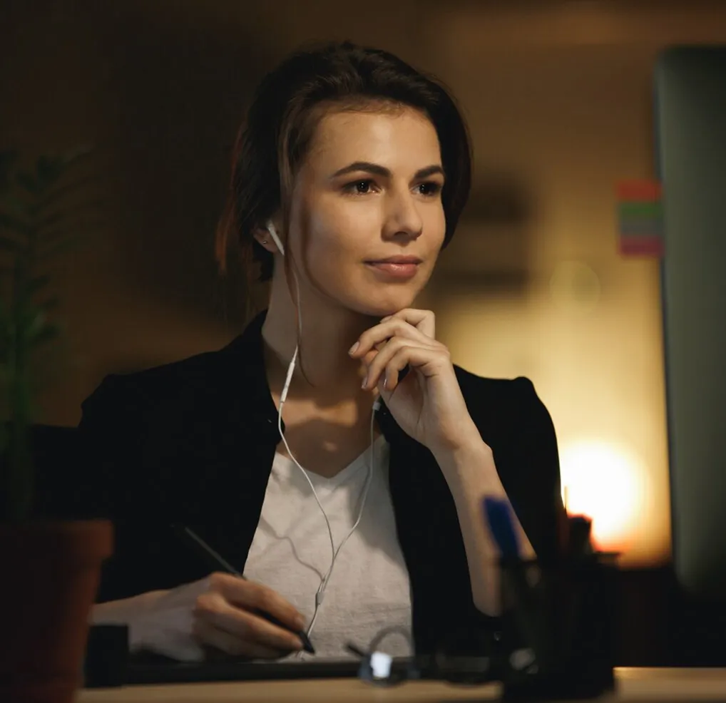 Woman wearing earphones, looking thoughtfully at a computer screen while holding a stylus pen.