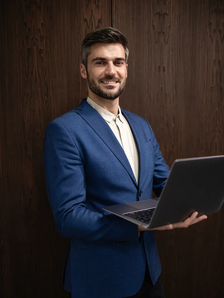 Smiling man wearing a blue blazer holding an open laptop against a dark wooden background.