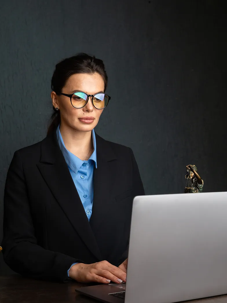 Woman with glasses in a black blazer and blue shirt working on a laptop against a dark background.
