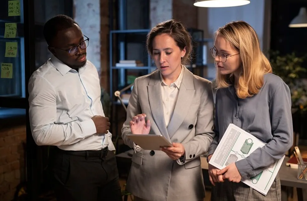 Three colleagues discussing work, one woman using a tablet, another holding documents, and a man listening attentively.