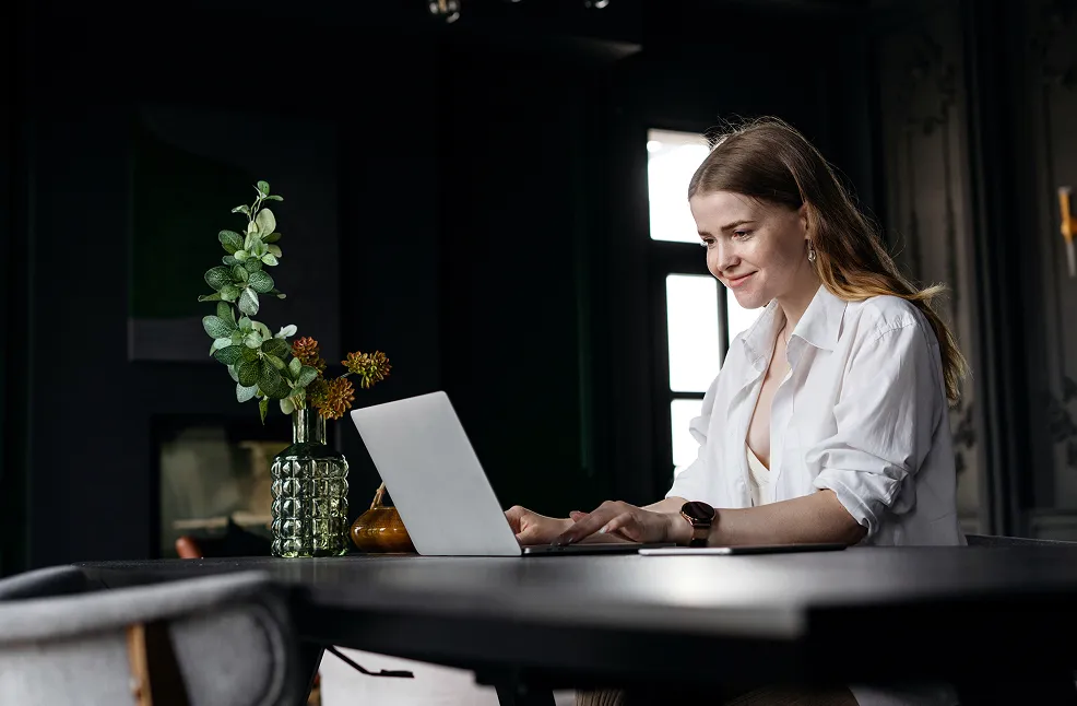 Young woman in a white shirt working on a laptop at a dark table with a vase of green and orange flowers nearby.