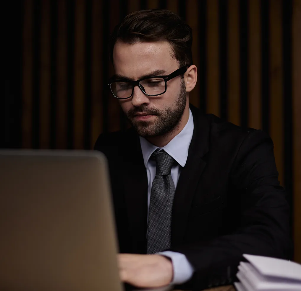 Man wearing glasses, a suit, and tie working on a laptop at a desk with stacked papers.