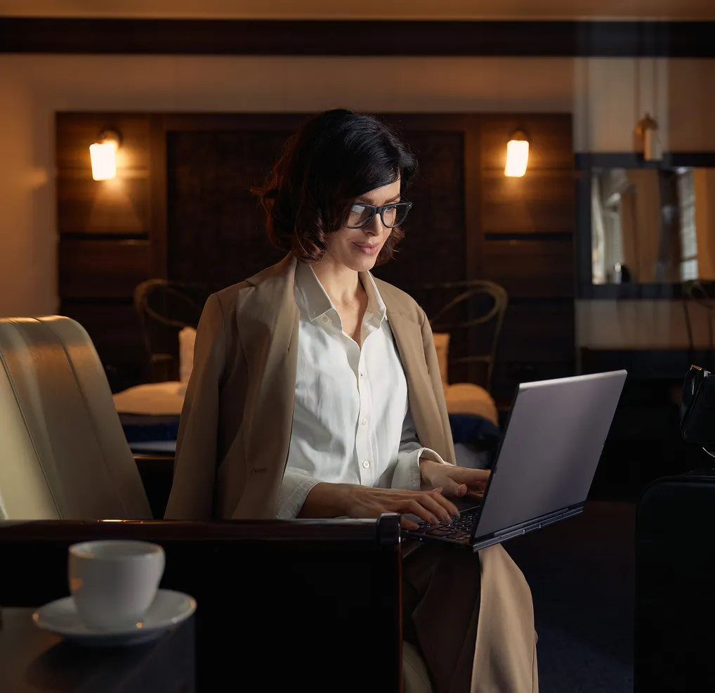 Woman in glasses and beige blazer working on a laptop in a dimly lit cozy room with warm wall lamps.