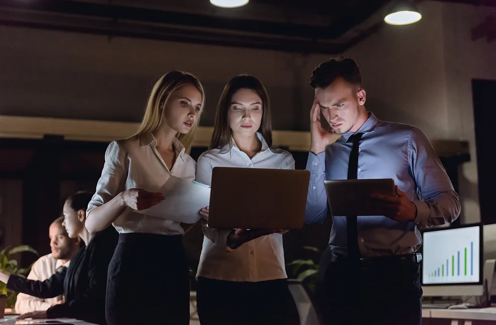 Three business colleagues working late, focused on a laptop and tablet in a dimly lit office.