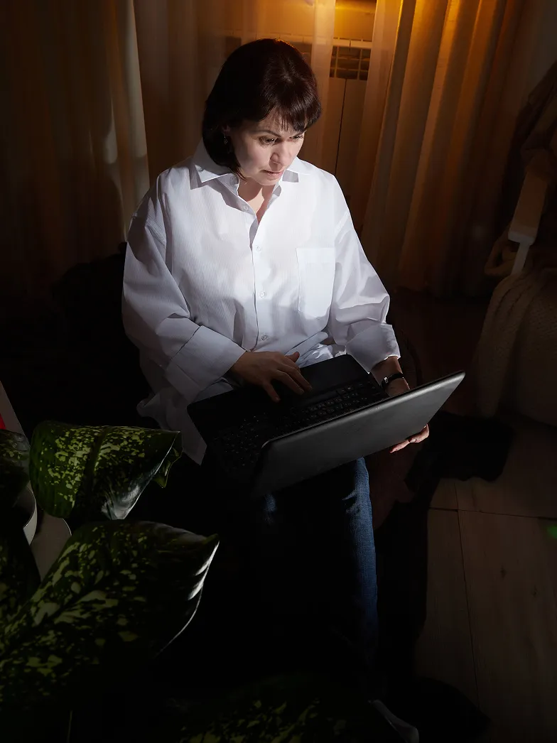 Woman in a white shirt working on a laptop in a dimly lit room with curtains and a plant nearby.