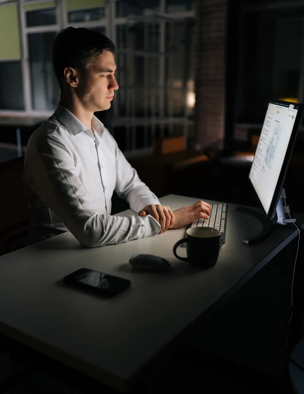 Man in a white shirt working on a desktop computer in a dark room with a mug, mouse, and smartphone on the desk.