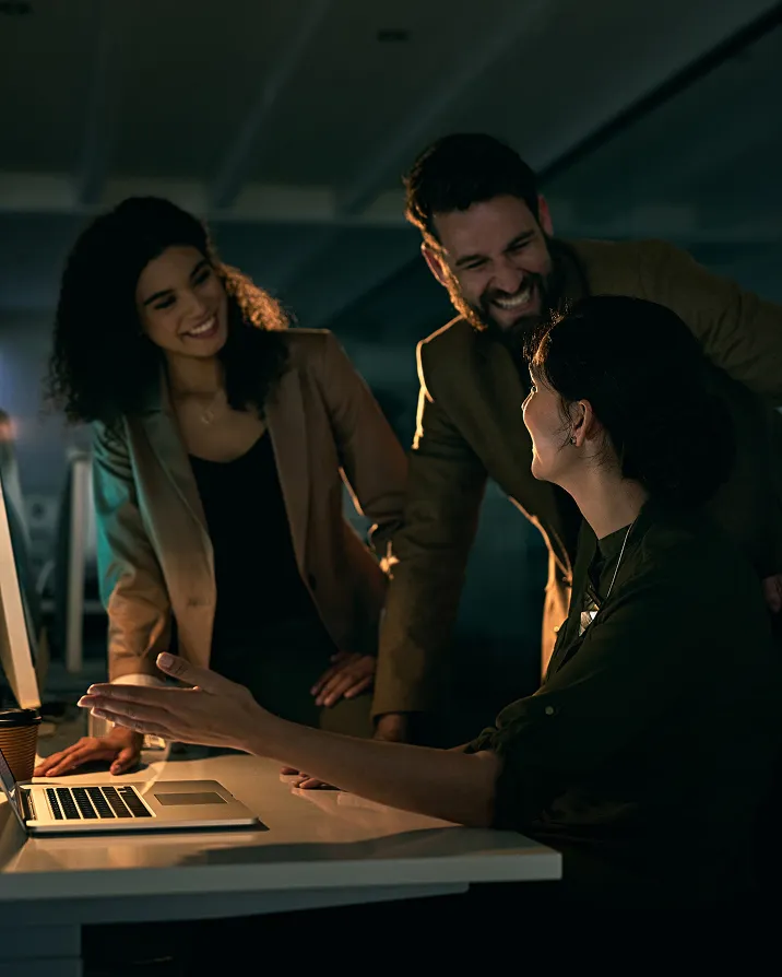 Three colleagues smiling and discussing work around a laptop in a dimly lit office.