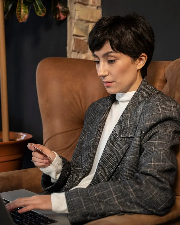 Woman in a gray plaid blazer sitting on a brown chair using a laptop and holding a card.