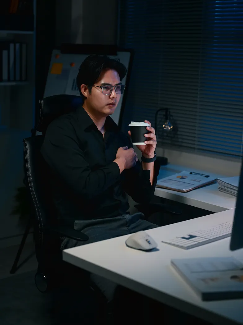 Man in glasses and black shirt sitting at a desk at night holding a cup, looking at a computer screen.