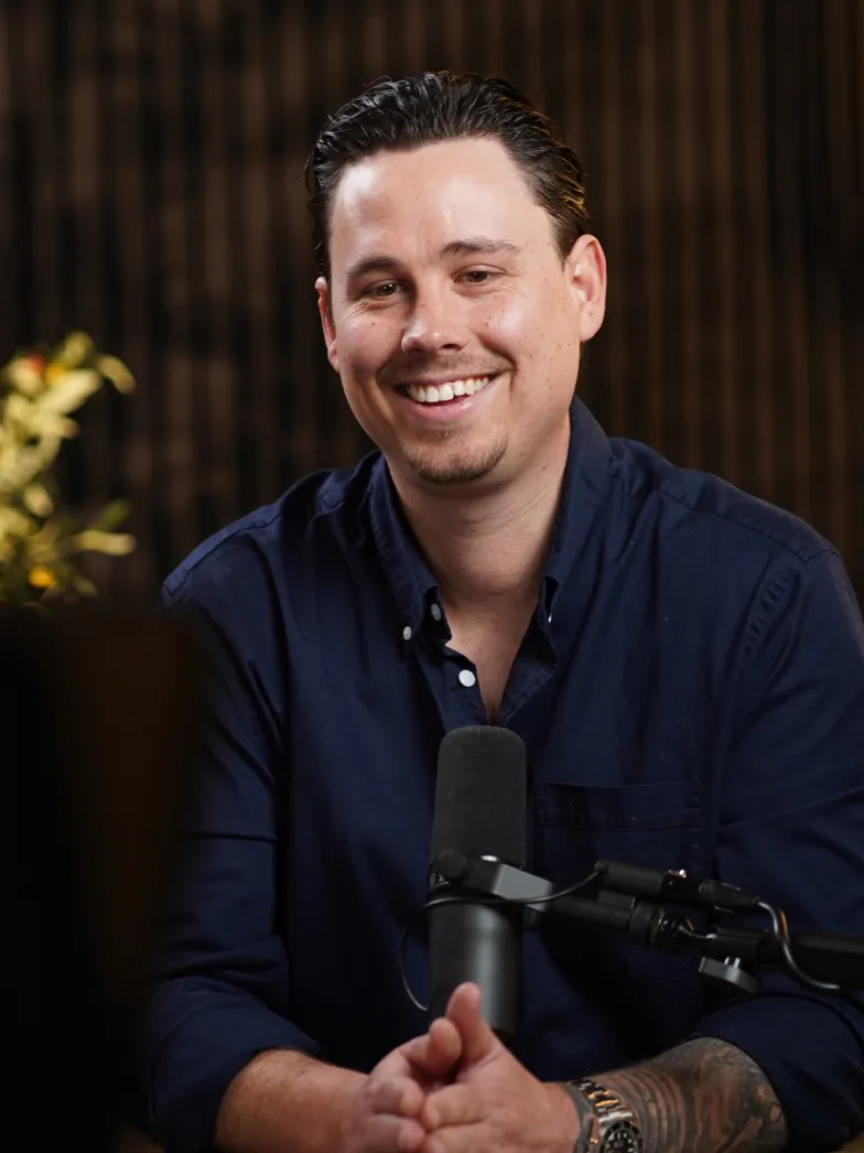 Smiling man with dark hair wearing a navy blue shirt sitting in front of a microphone and clasping his hands.