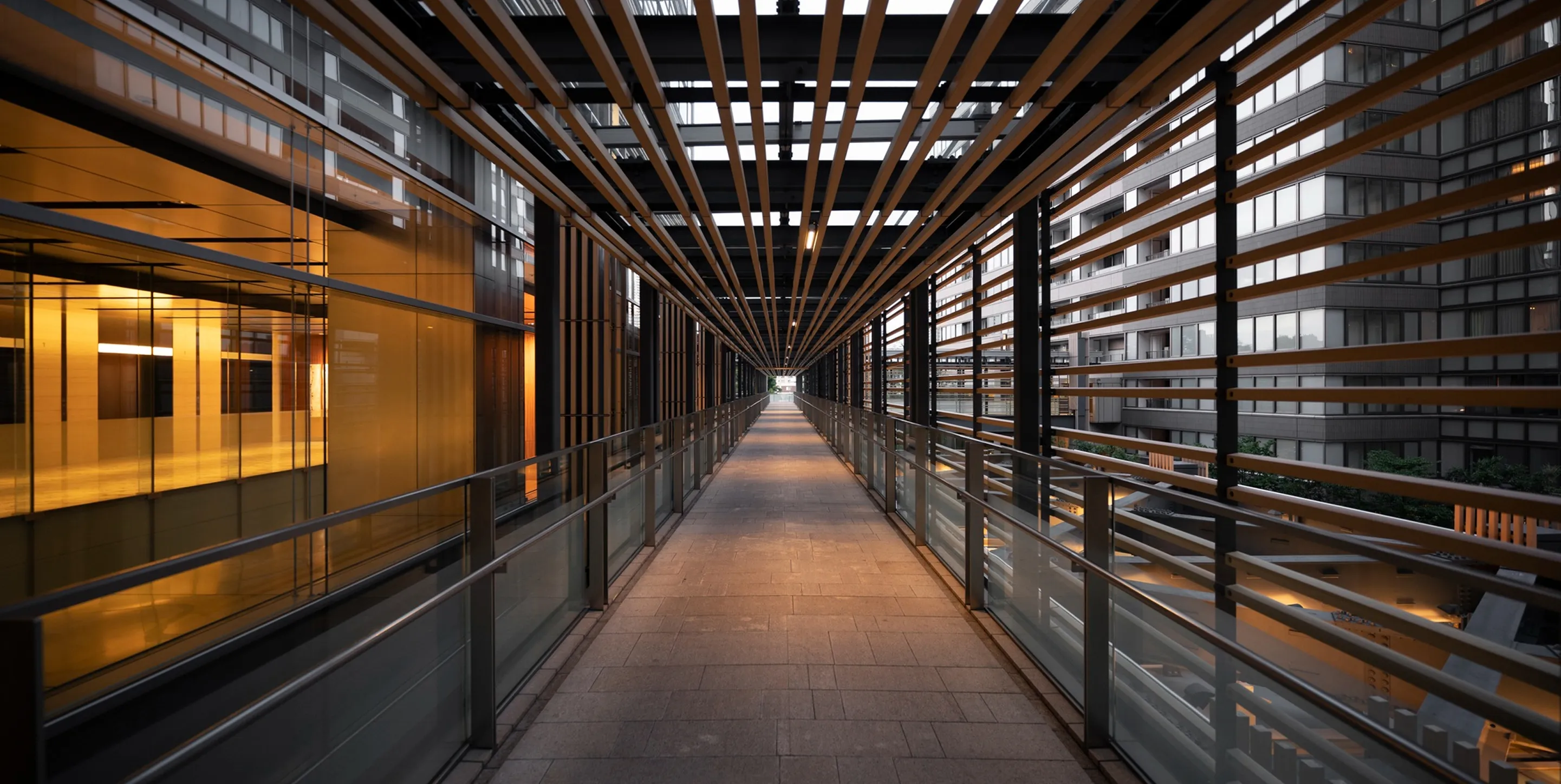 Modern covered walkway between buildings with glass railings and horizontal wooden slats overhead and to the sides.