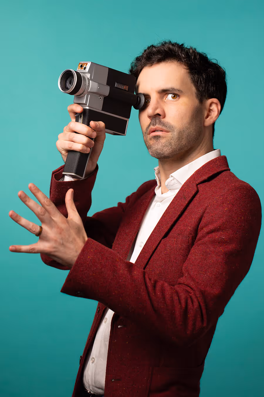Man in a red blazer looking through a vintage film camera against a teal background.