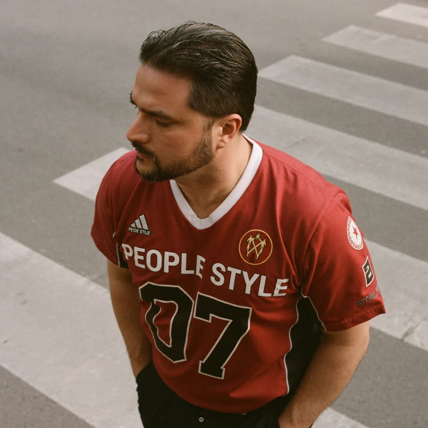 Man in a red jersey standing on a crosswalk, looking down, with his hands in his pockets