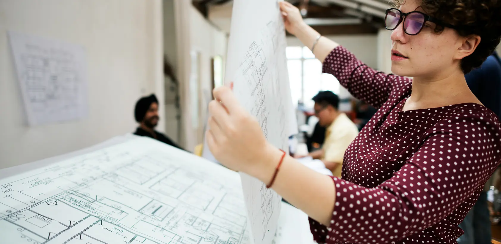 Woman wearing glasses and a red patterned top examining architectural blueprints in an office with colleagues in the background.