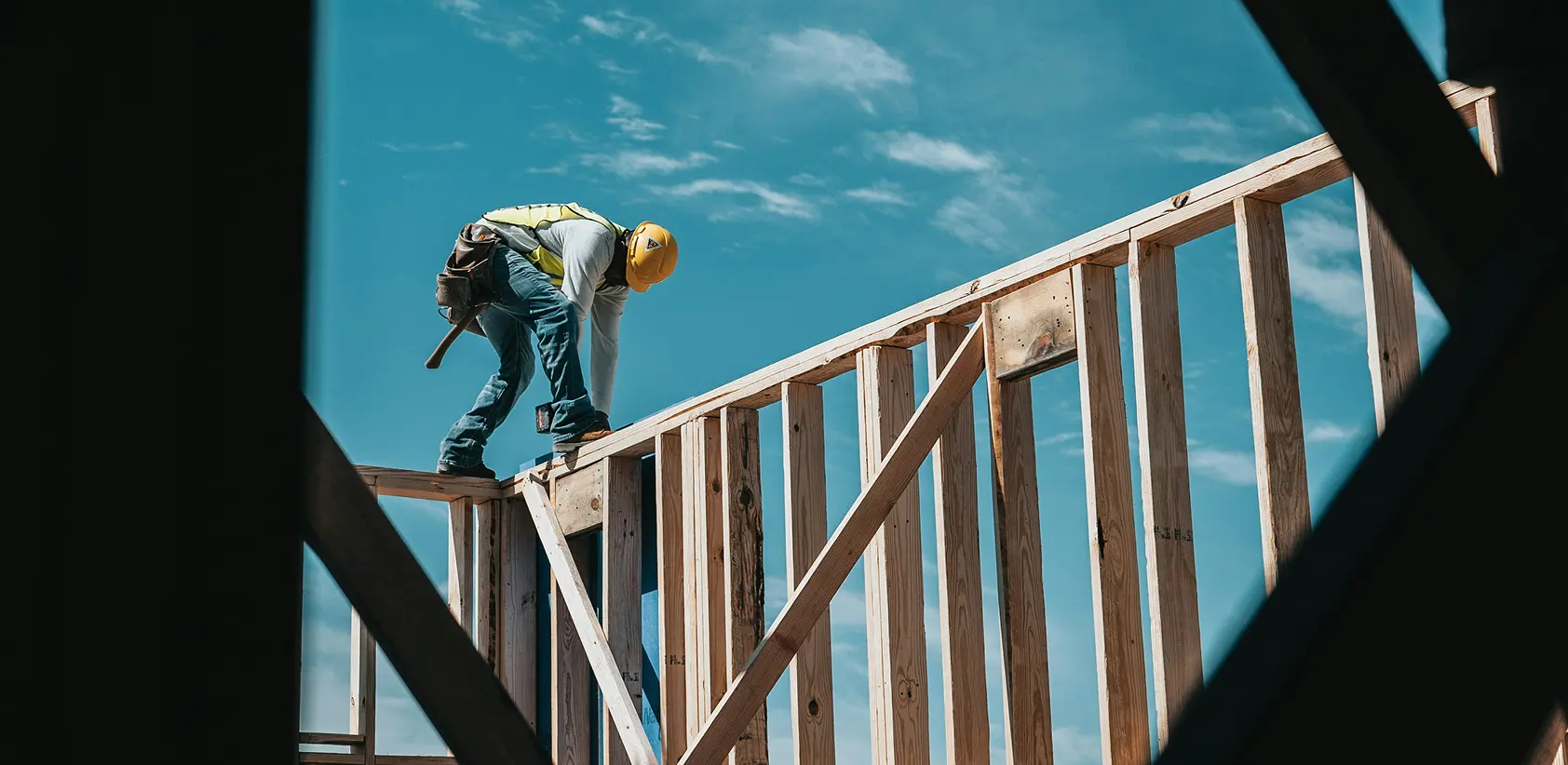Construction worker wearing a yellow helmet and safety vest working on framing a wooden building under a blue sky.