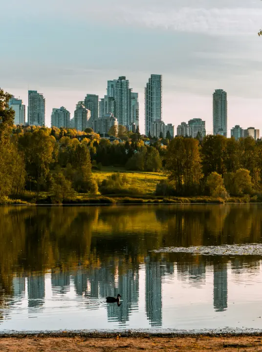 City skyline with tall buildings reflected in a calm lake surrounded by trees and greenery.