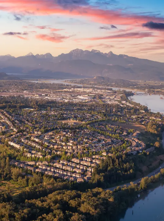 Aerial view of a suburban neighborhood with winding roads, dense trees, a river, and mountains under a colorful sunset sky.