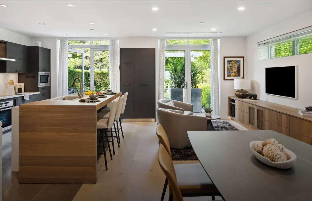 Modern open kitchen and living room with wooden island, bar stools, beige chairs, large windows, and a TV mounted on the wall.