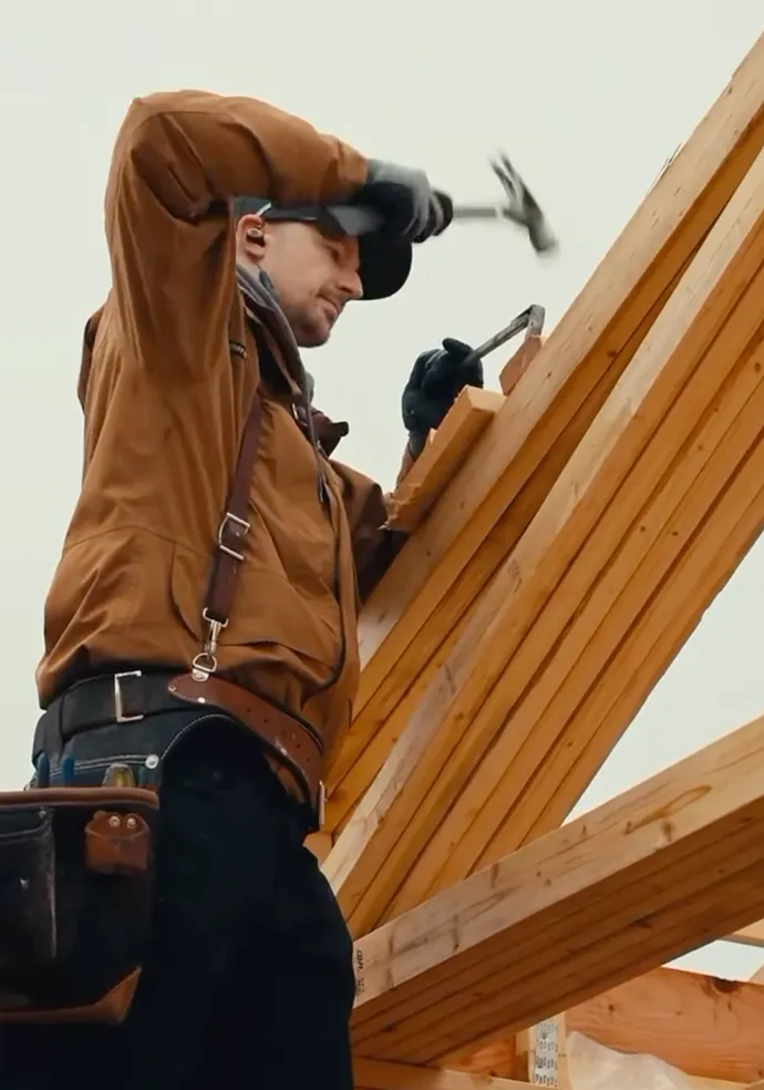 Carpenter wearing a brown jacket and gloves hammering a nail into a wooden beam.