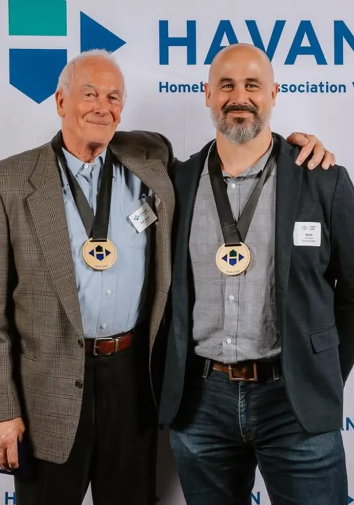 Two men wearing medals around their necks standing in front of a Havan backdrop; the older man on the left wears a plaid blazer and blue shirt, the younger man on the right wears a dark blazer and gray shirt.