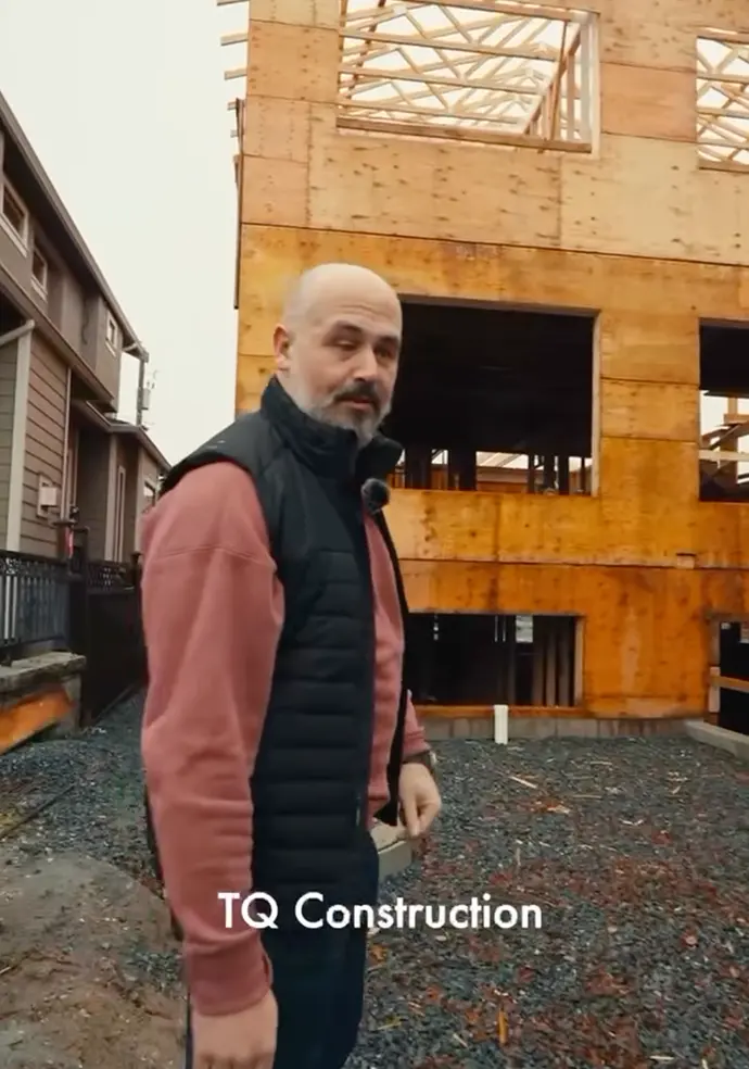 Man in a black vest and pink sweatshirt standing at a construction site with a partially built wooden house behind him.
