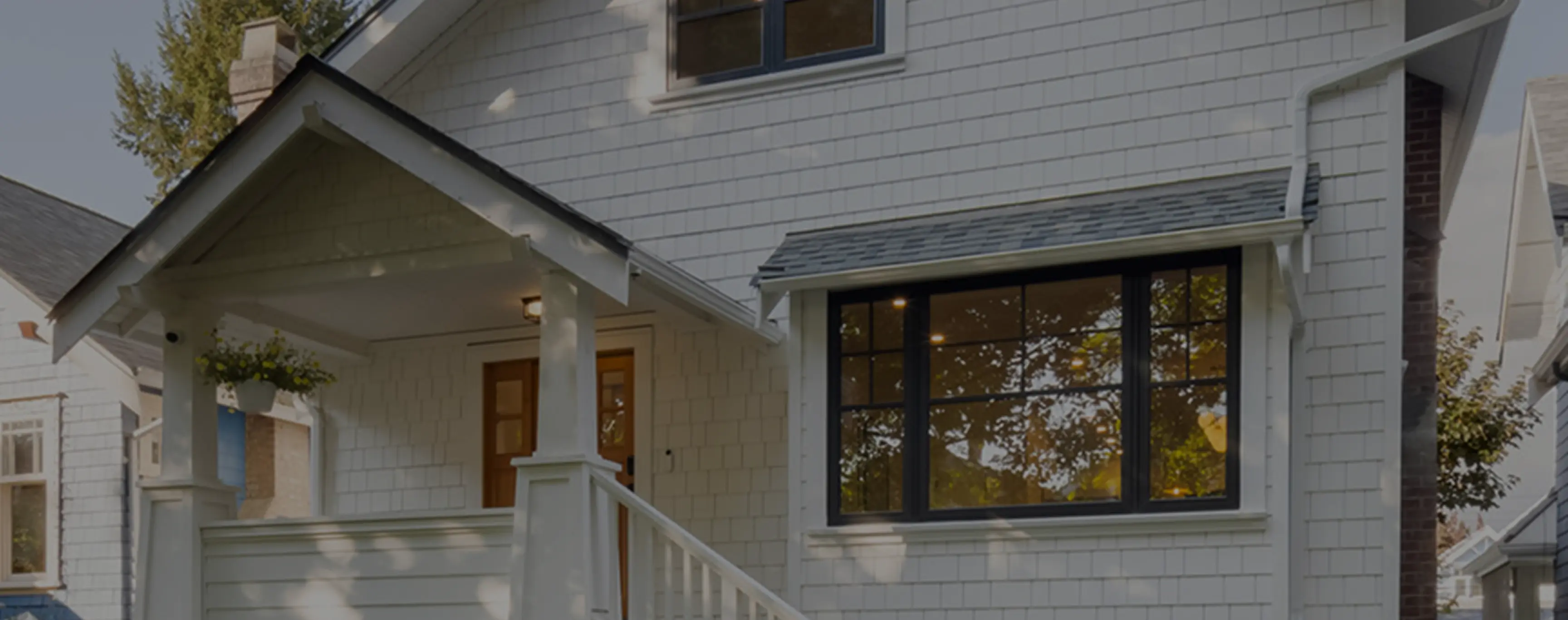 White house exterior with a porch, wooden front door, large black-framed window, and hanging flower pot.