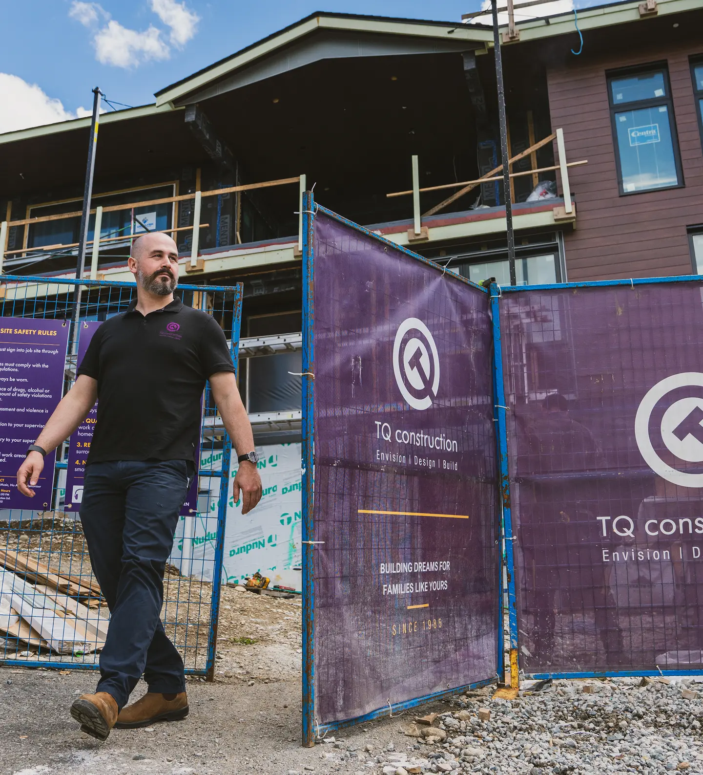 Man in black polo shirt and brown boots walking out of a construction site with purple TQ construction safety fences around a partially built house.