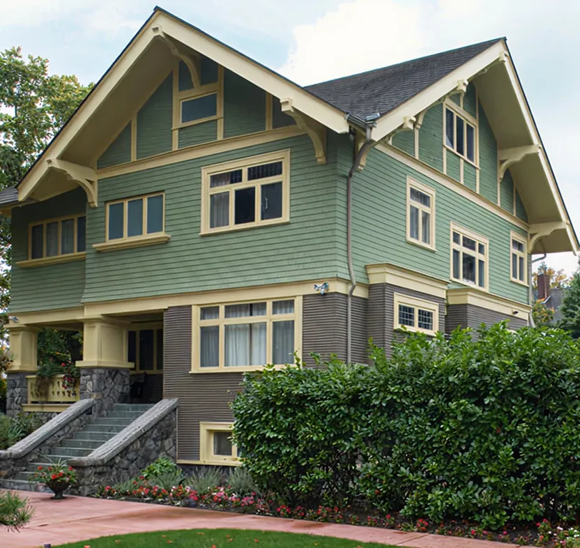 Two-story craftsman-style house with green and beige siding, stone steps, and a well-maintained garden with shrubs and flowers.