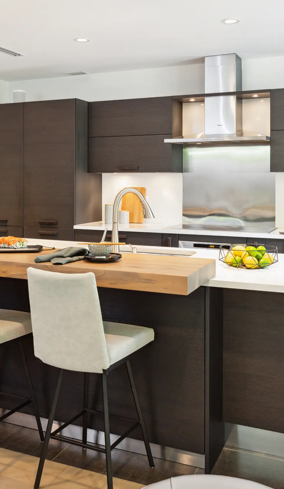 Modern kitchen with dark wood cabinets, a wooden countertop bar with beige chairs, and a stainless steel range hood above the stove.