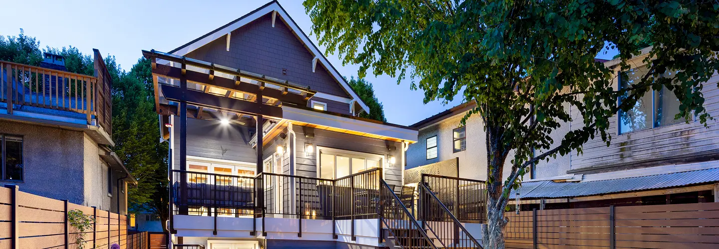 Modern gray two-story house with lit porch, metal railing stairs, and surrounding wooden fence at dusk.