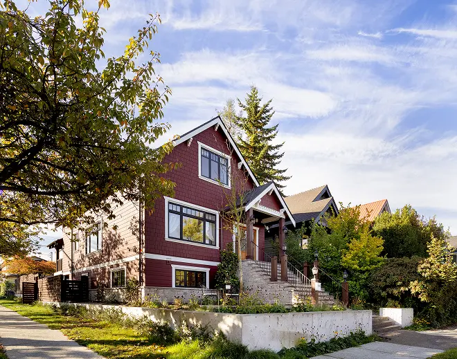 A red two-story house with white trim and a front staircase surrounded by trees and greenery under a partly cloudy sky.