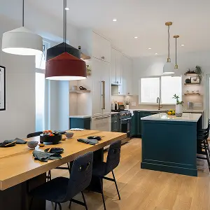 Modern kitchen with navy blue island, wooden dining table with black chairs, and pendant lights above.