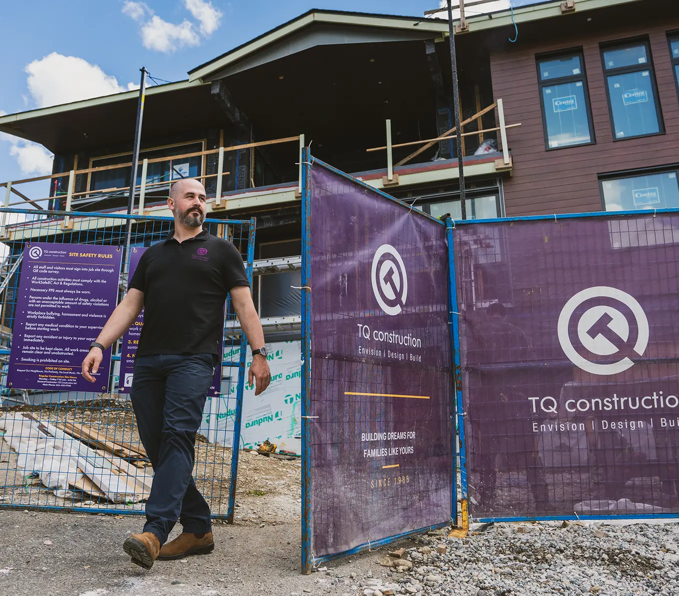 Man in black polo shirt and boots walking away from a construction site with purple TQ Construction banners on a metal fence.