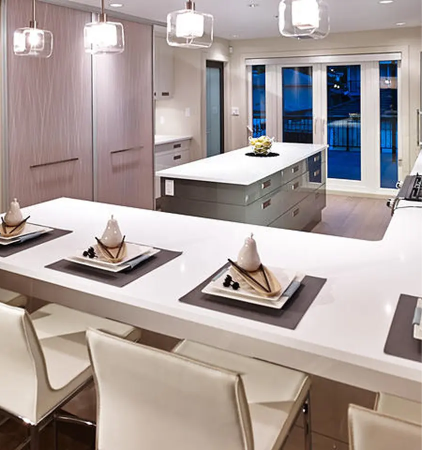 Modern kitchen with white countertops, three pendant lights, cream-colored chairs, and neatly set place settings with napkins and decorative pears.