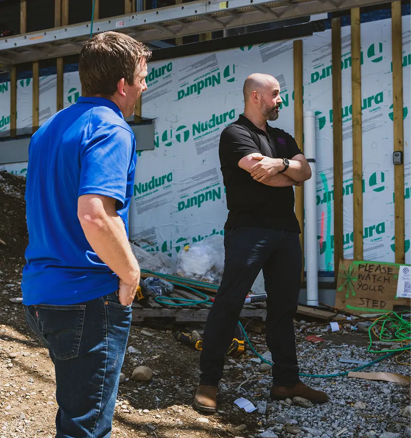 Two men discussing at a construction site with exposed wooden framing and a sign that says 'Please Watch Your Step'.