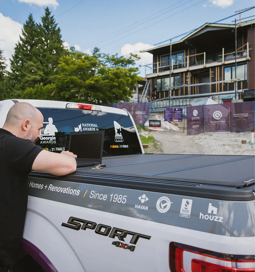 Man leaning on the back of a pickup truck with a laptop open, with a construction site and partially built house in the background.